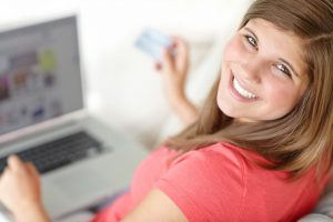 Una chica mira a cámara mientras está con un ordenador portátil en las rodillas y sostiene una tarjeta de crédito en la mano derecha se ve que acaba de entrar en una página de una clínica dental gracias una campaña de publicidad SEM realizada por Medicalta, marketing sanitario para clínicas.